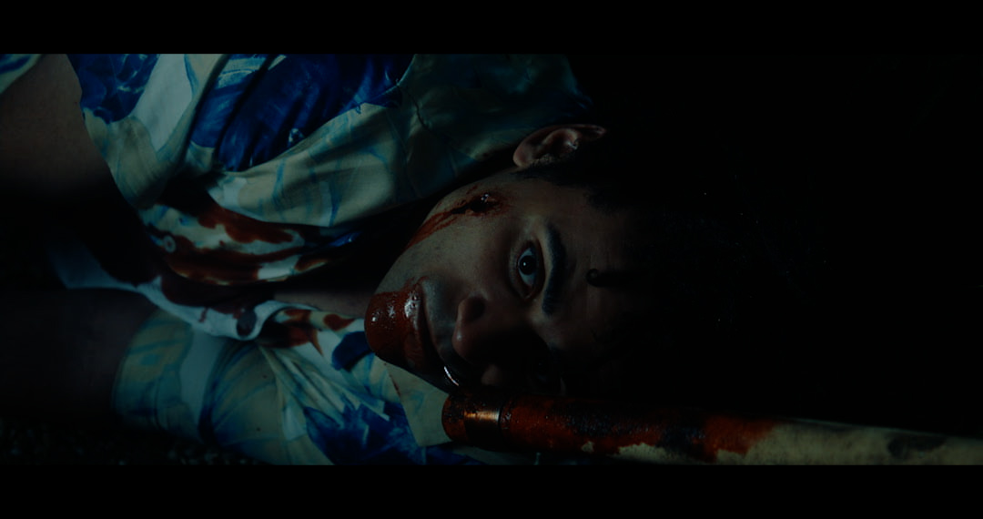 Close up of the bloodied man's face revealing a large, round, wound on the left side of his head and blood pooling around his mouth